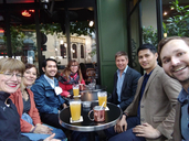 Seven people sitting outside by a couple of small tables. They are having drinks and looking towards the camera, smiling.
